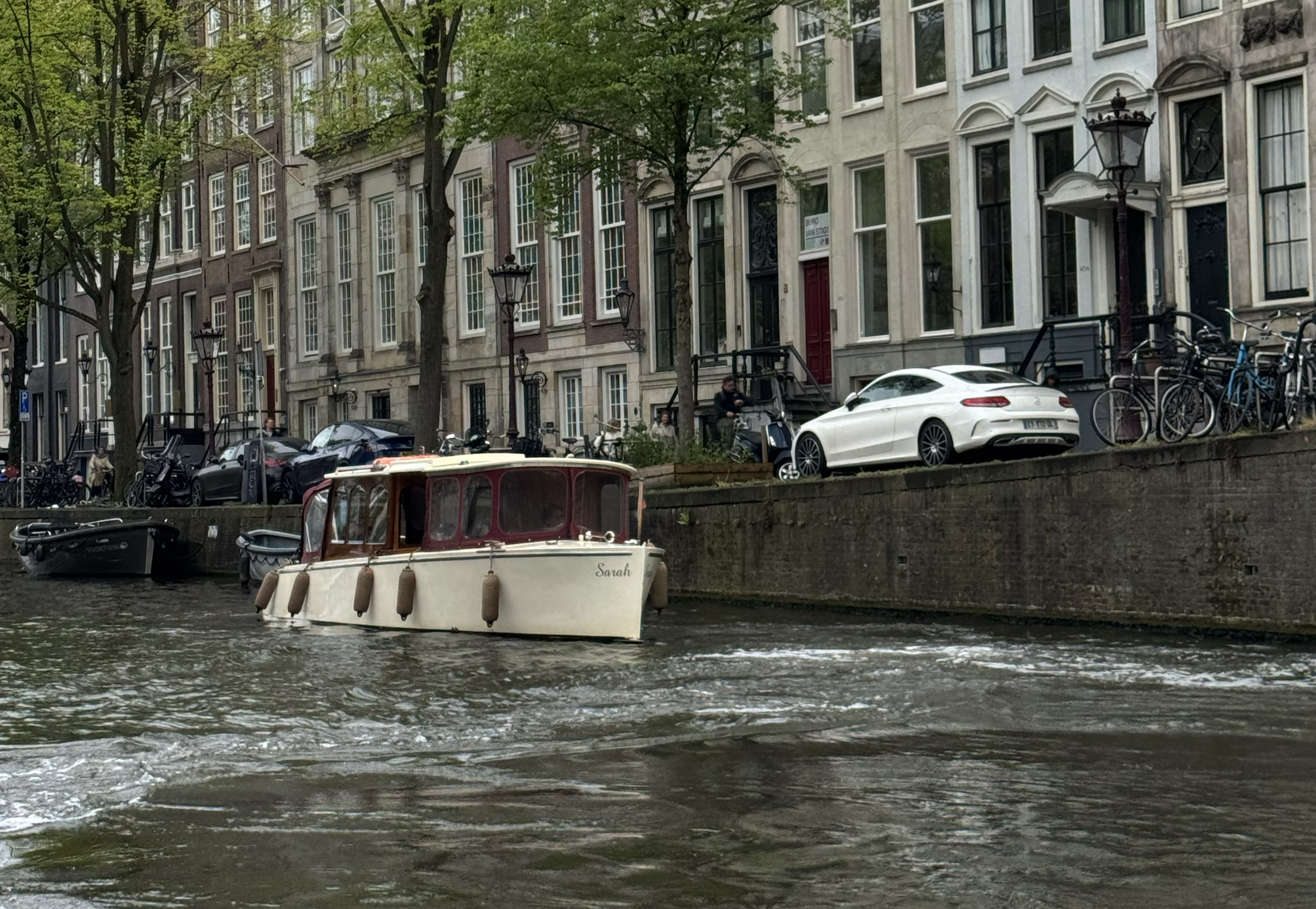 A boat floats on a canal, with tall buildings and trees in the background.