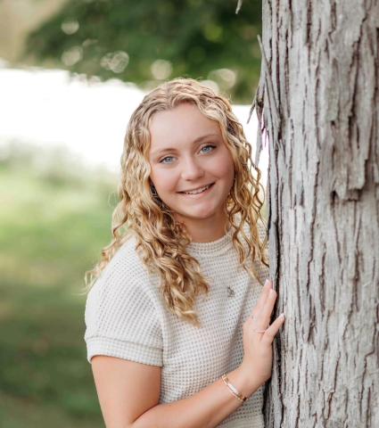 Ella faces the camera with a bright smile as she leans against a tree.