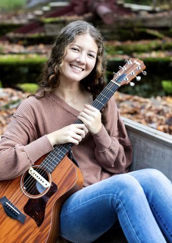 Taylor Stephens is pictured holding a brown guitar wearing a brown long sleeve and jeans. Fallen leaves shadow the ground behind her.