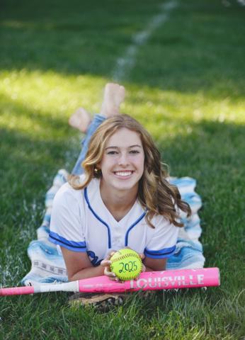 Emily Pratt lays on her stomach in her softball uniform with a bat and a softball in front of her.