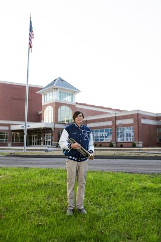 Jack Hood standing in front of Trinity High School. He is wearing his letterman jacket and holding a trumpet in his hands.