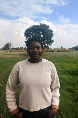 Matsaudza is pictured smiling in a field with a beautiful tree in the background. She is wearing a cream long-sleeve with her hands resting in her pockets.