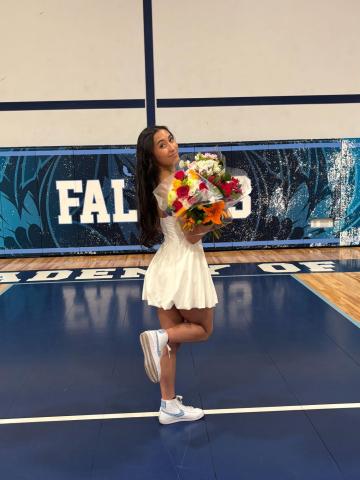 Anastasia Curitti stands facing away with her right knee bent back and looks over her shoulder. She carries a large bouquet of colorful flowers over her right shoulder and wears a white dress. She is in a gymnasium with blue floors.