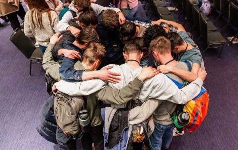 A group of boys huddles together with their hands on each other's shoulders and heads close. 