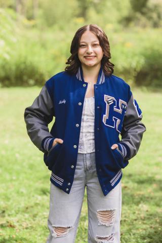Madilyn Paterno standing in a green field, wearing a blue letterman jacket with a white t-shirt and jeans, smiling at the camera