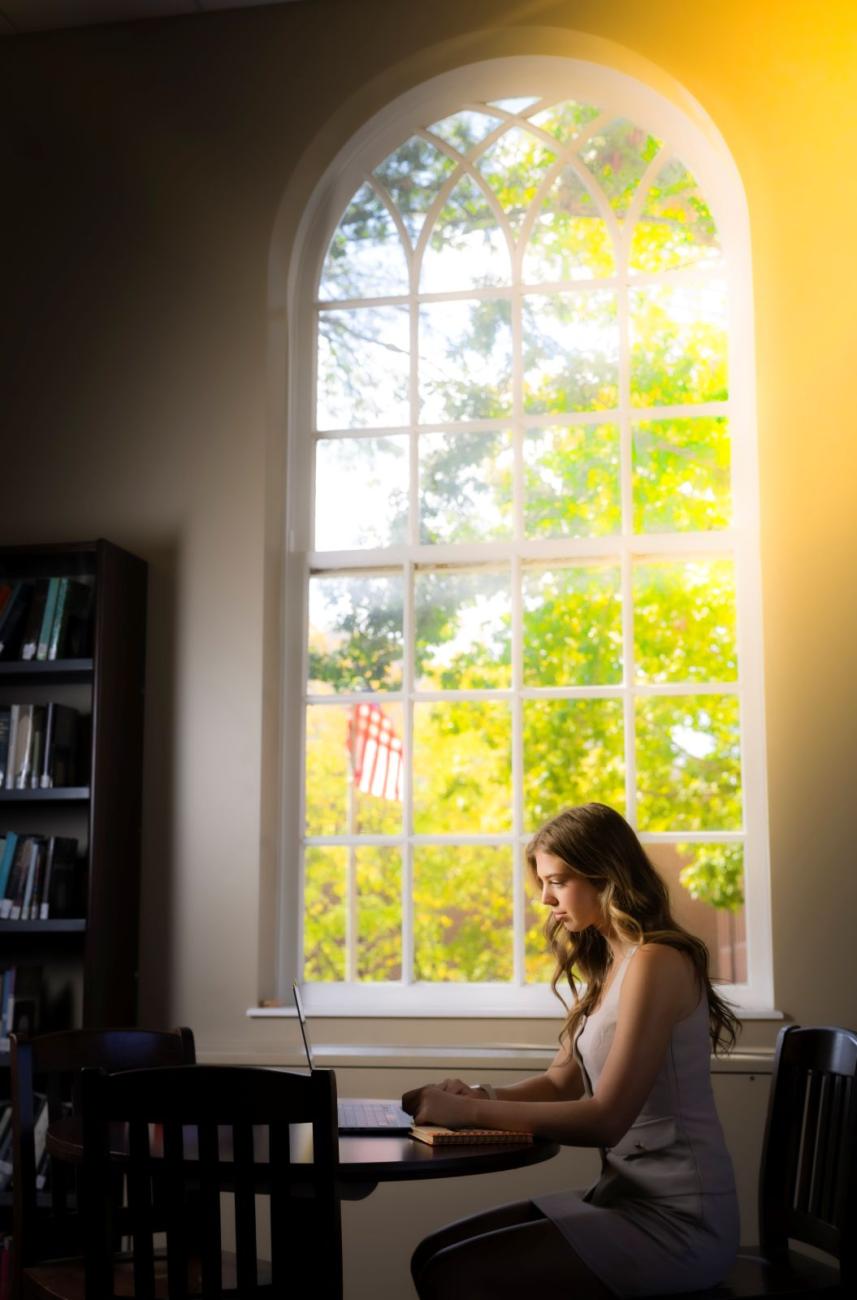 Stover Scholar sits by a window in the library with the sun streaming through