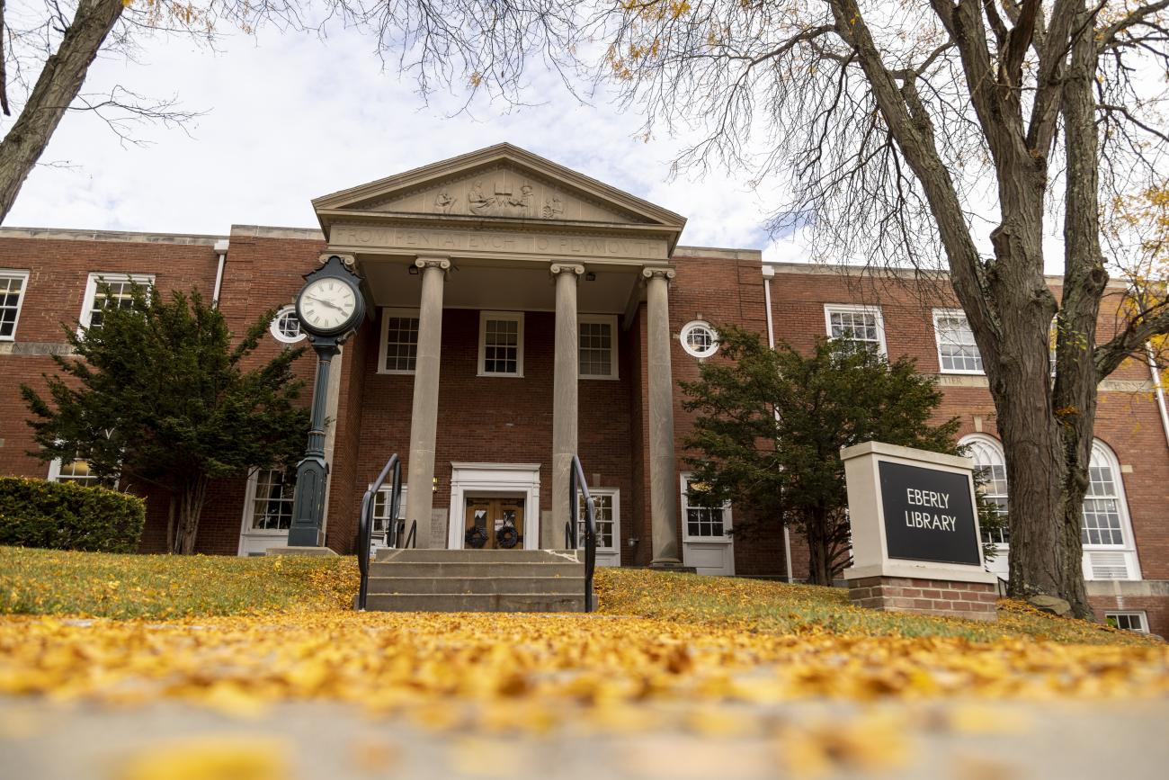 Fallen yellow leaves cover the sidewalk in front of Eberly Library in autumn; the library stands tall against a grey sky with bare trees and evergreen shrubs in front of it.
