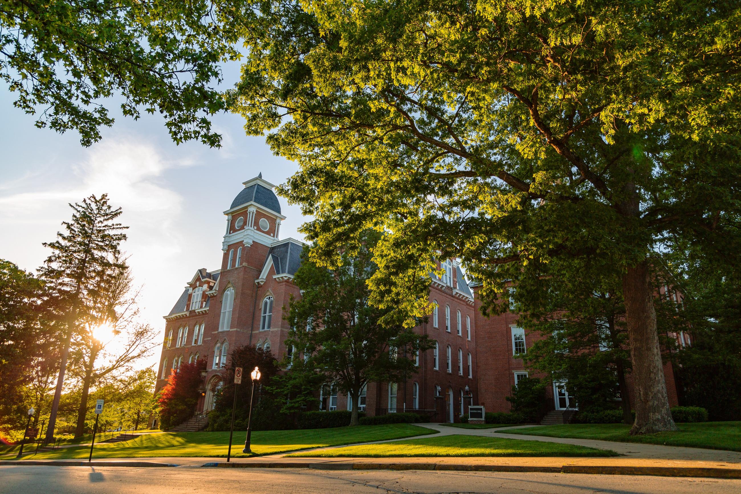Thick green trees stand in front of Miller and Hanna Halls as the sun's rays peak through the leaves. 