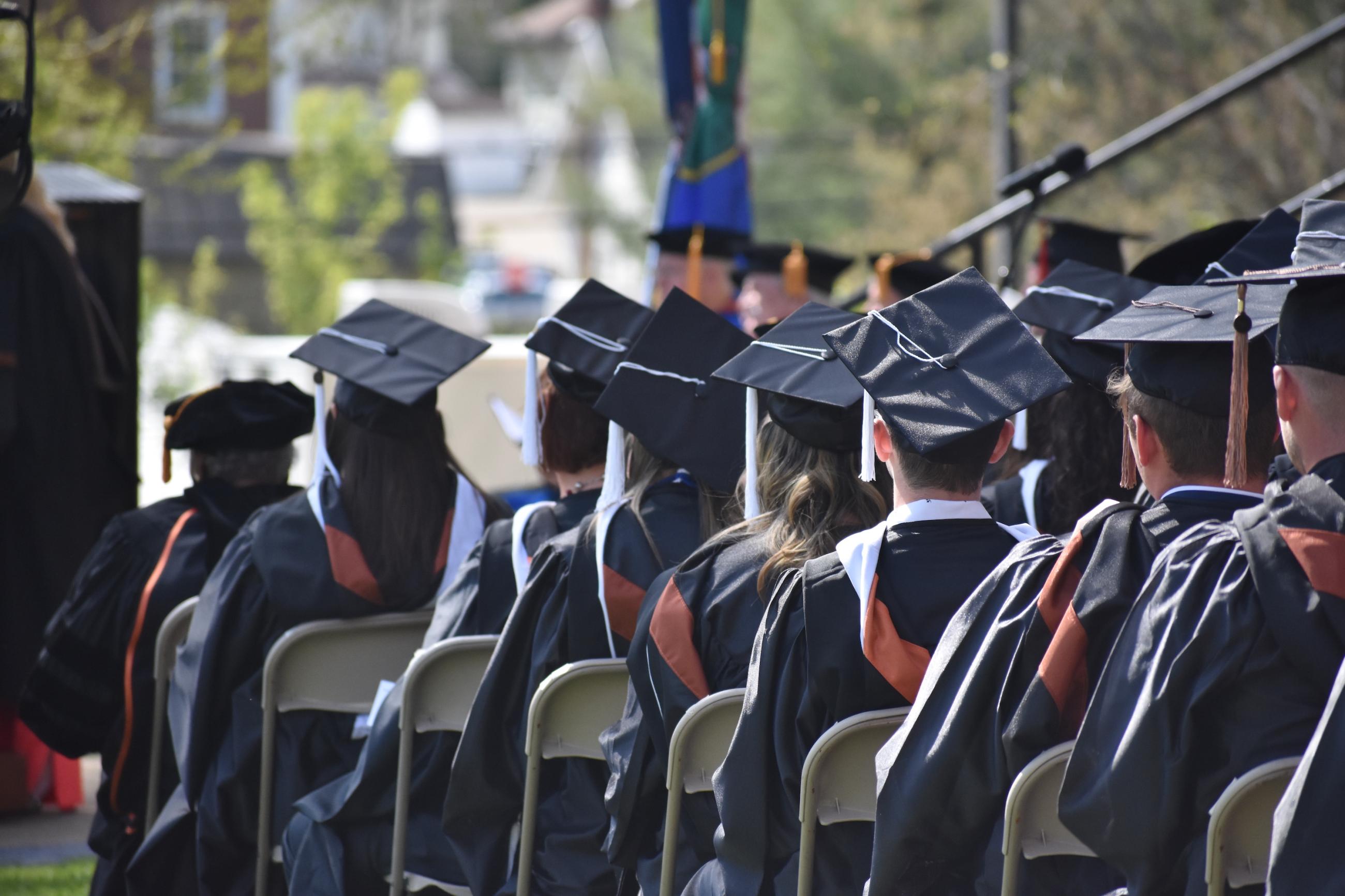 Waynesburg University graduates sit in their caps and gowns on the lawn of Miller Hall with their backs to the camera.