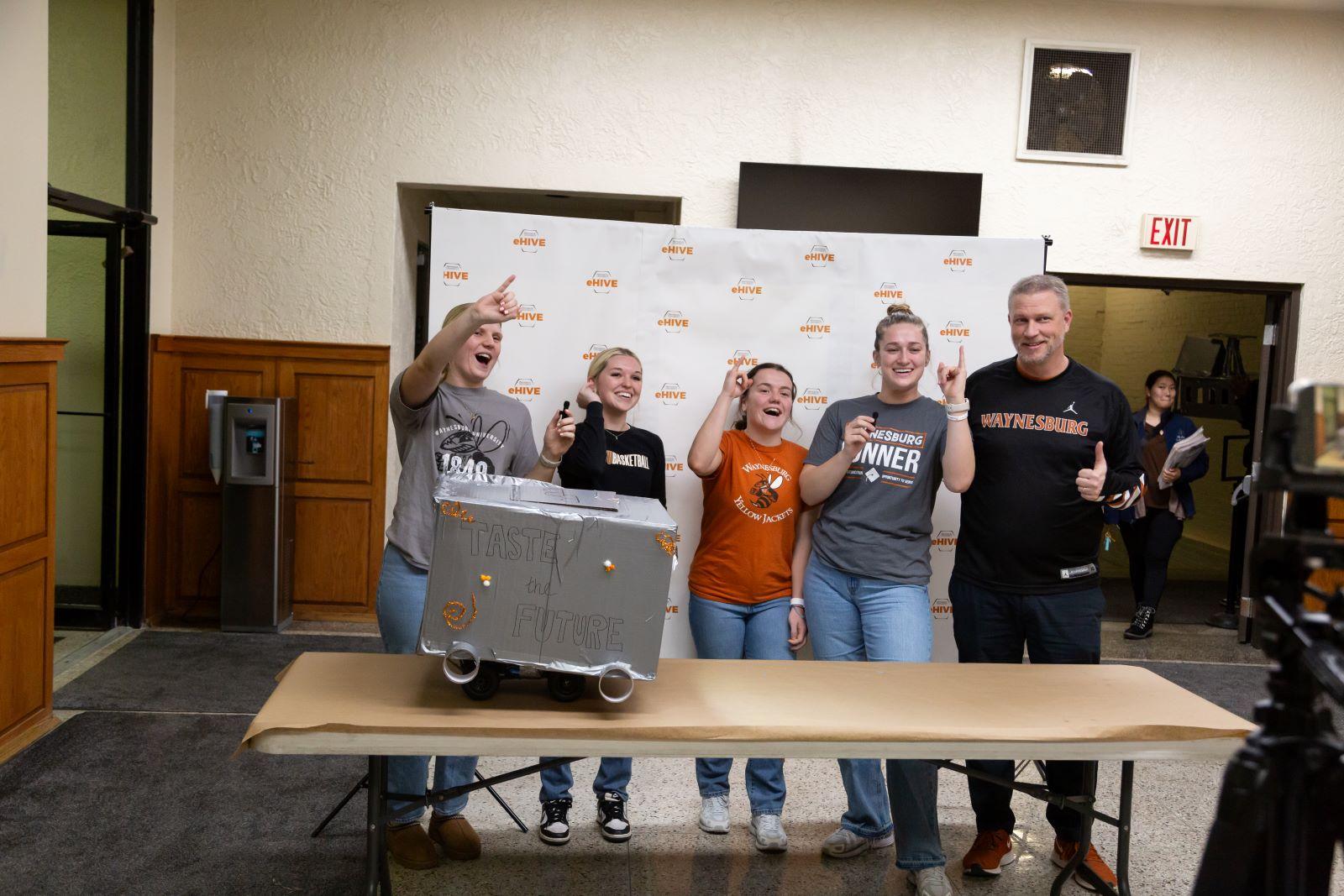 A group of five hold their pinky fingers up to the camera with excitement as their final product sits in front of them