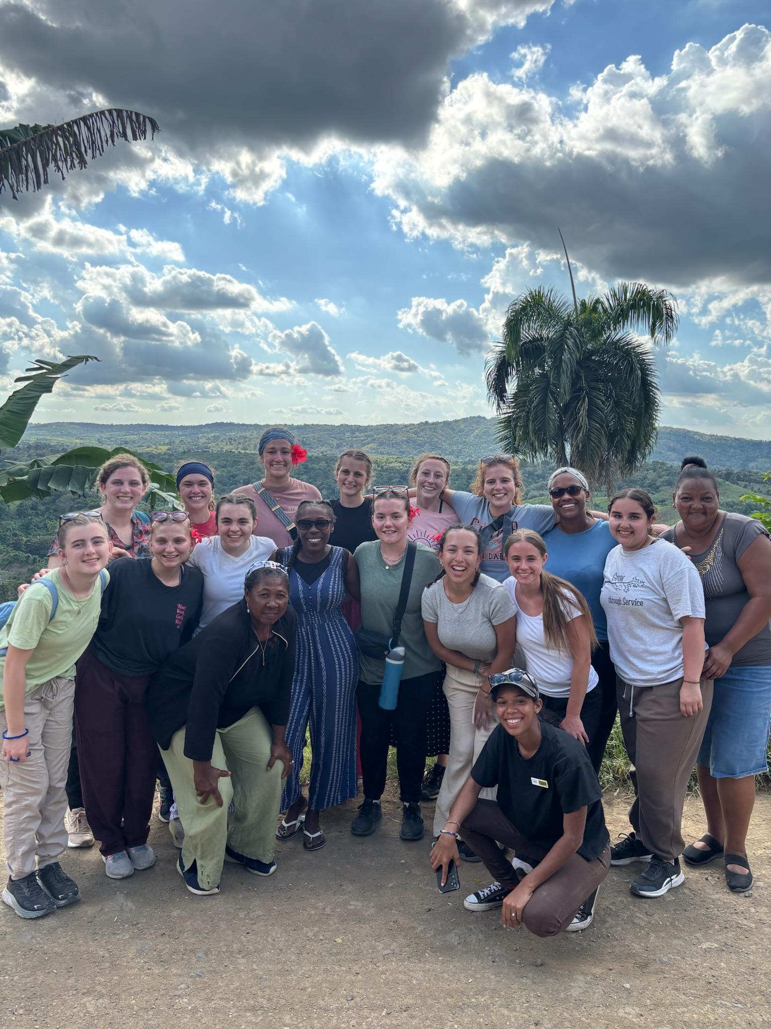 The group's women stand in front of the green mountain for a group photo. A palm tree sits behind them in their brightly color clothes. 