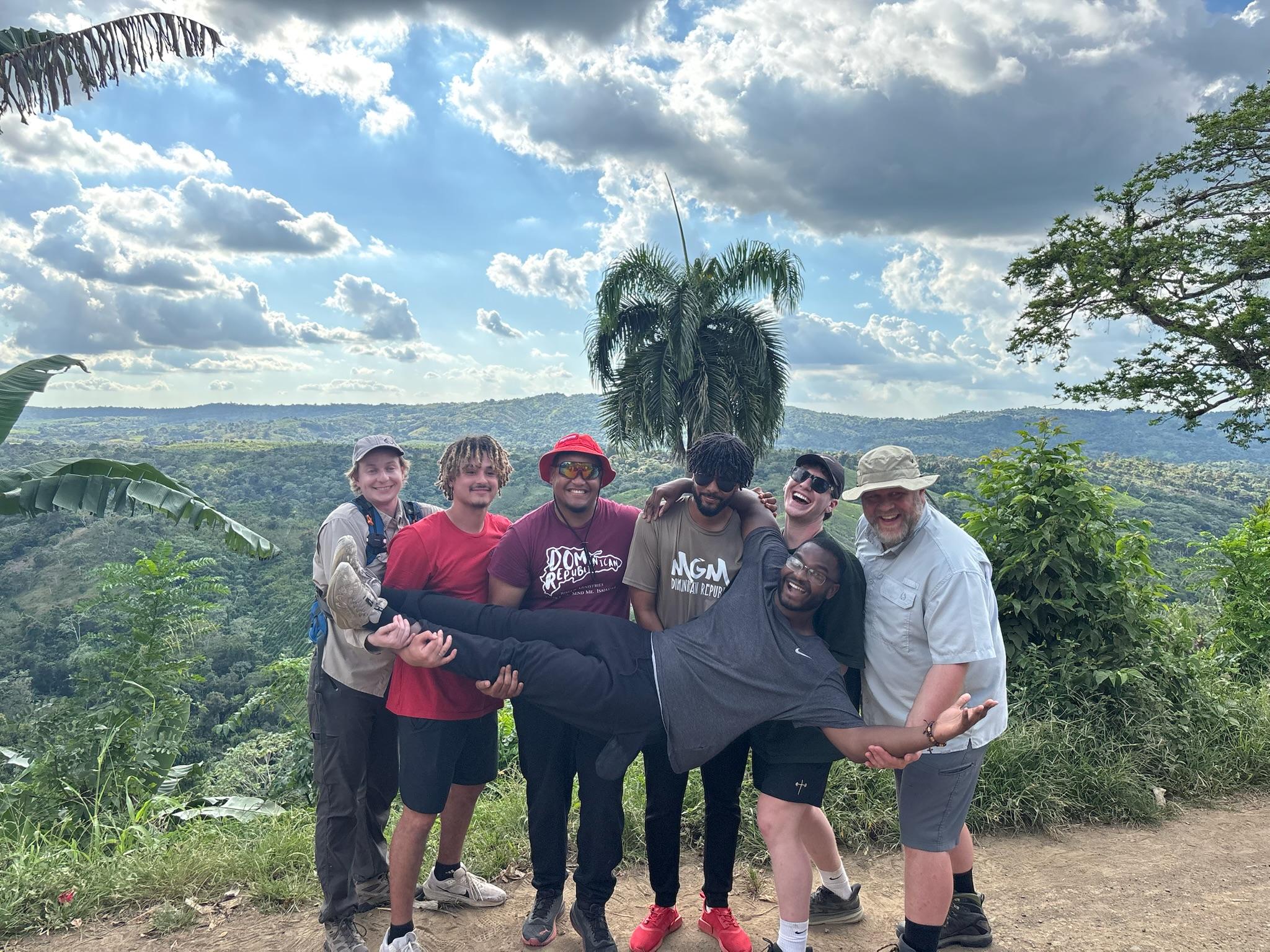 The group's men stand in front of the green mountain with one student held in front of them. Each one is smiling or laughing joyfully. 