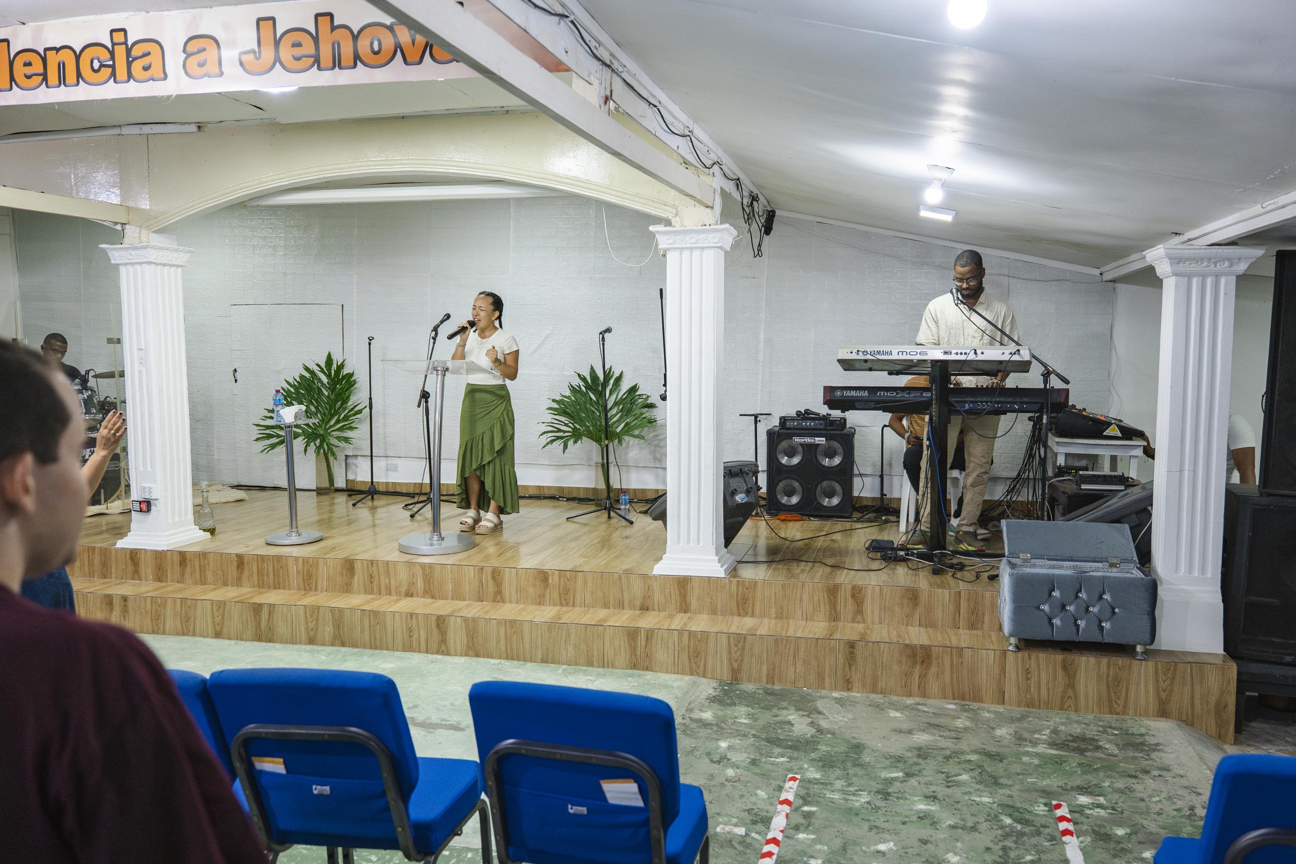 Two Waynesburg students worship together. One wearing a green skirt and a white shirt sings as she holds the microphone while  the other wearing a white shirt plays the piano nearby. 