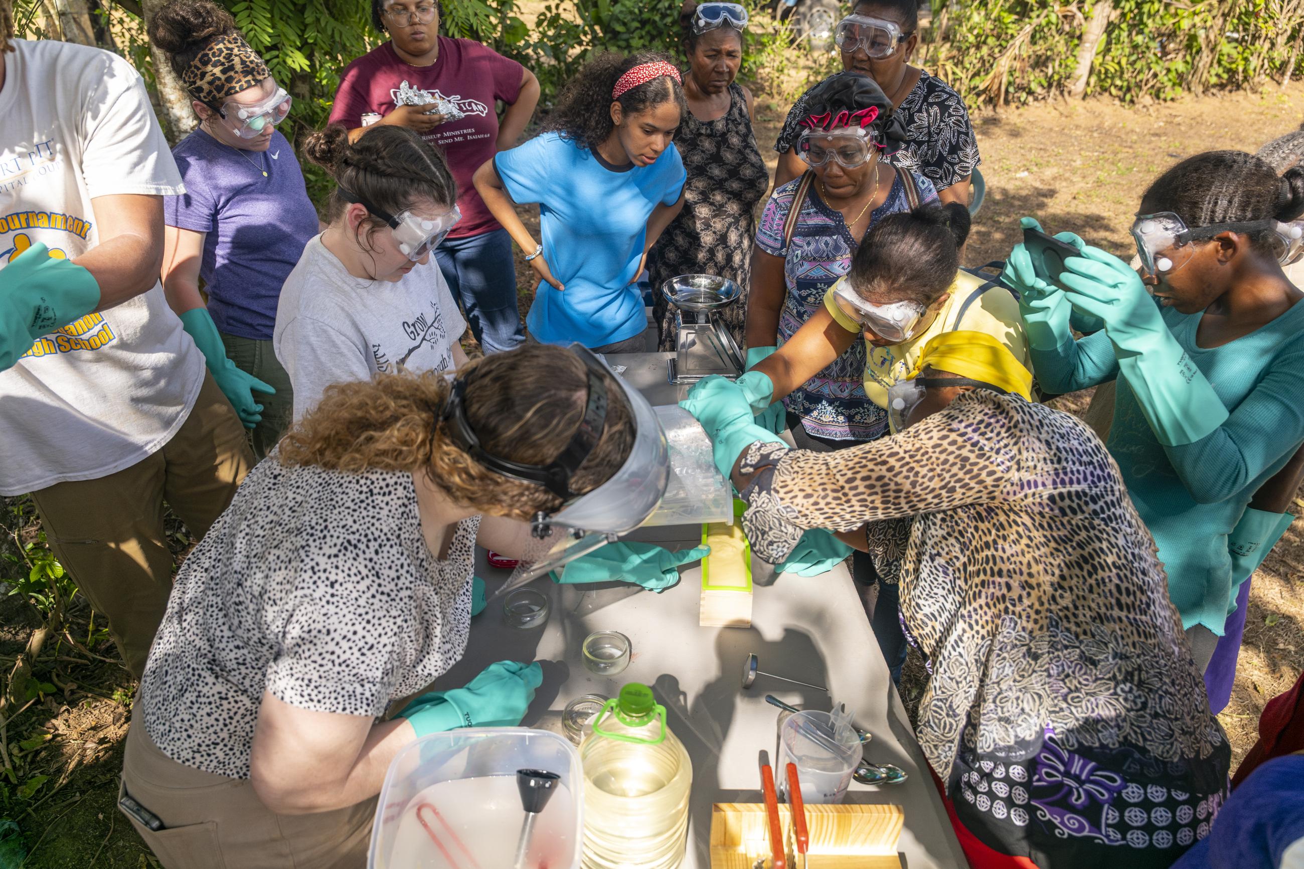 Multiple women with bright green gloves and masks help make soap using buckets.