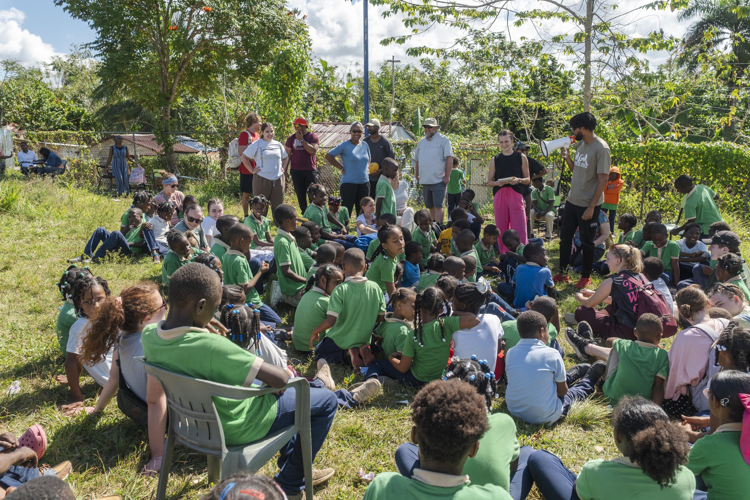 Kids sit around two people while one speaks into a megaphone. The other holds a notebook in her hands. 