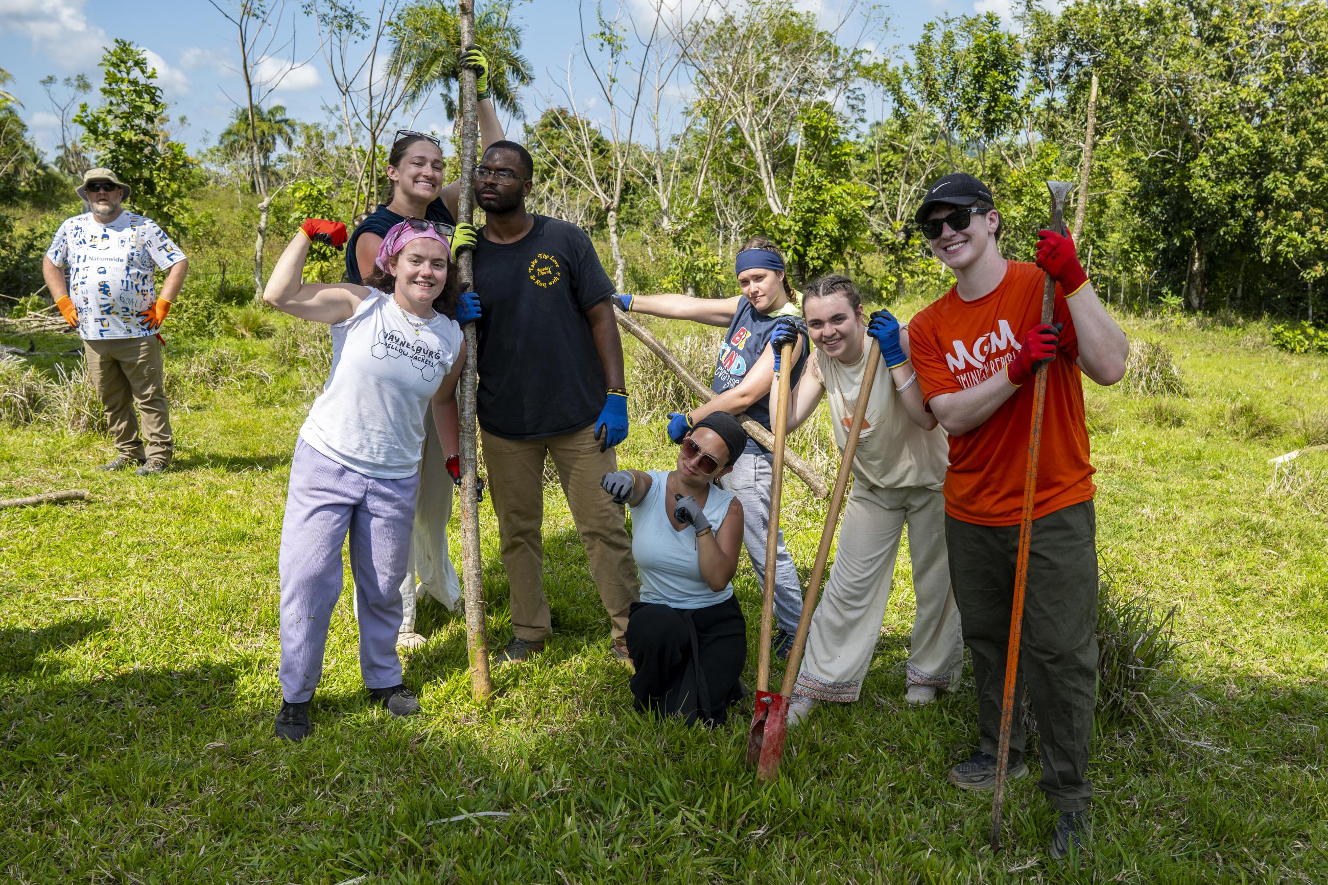 Seven students pose for the camera with branches and digging materials in their hands. 