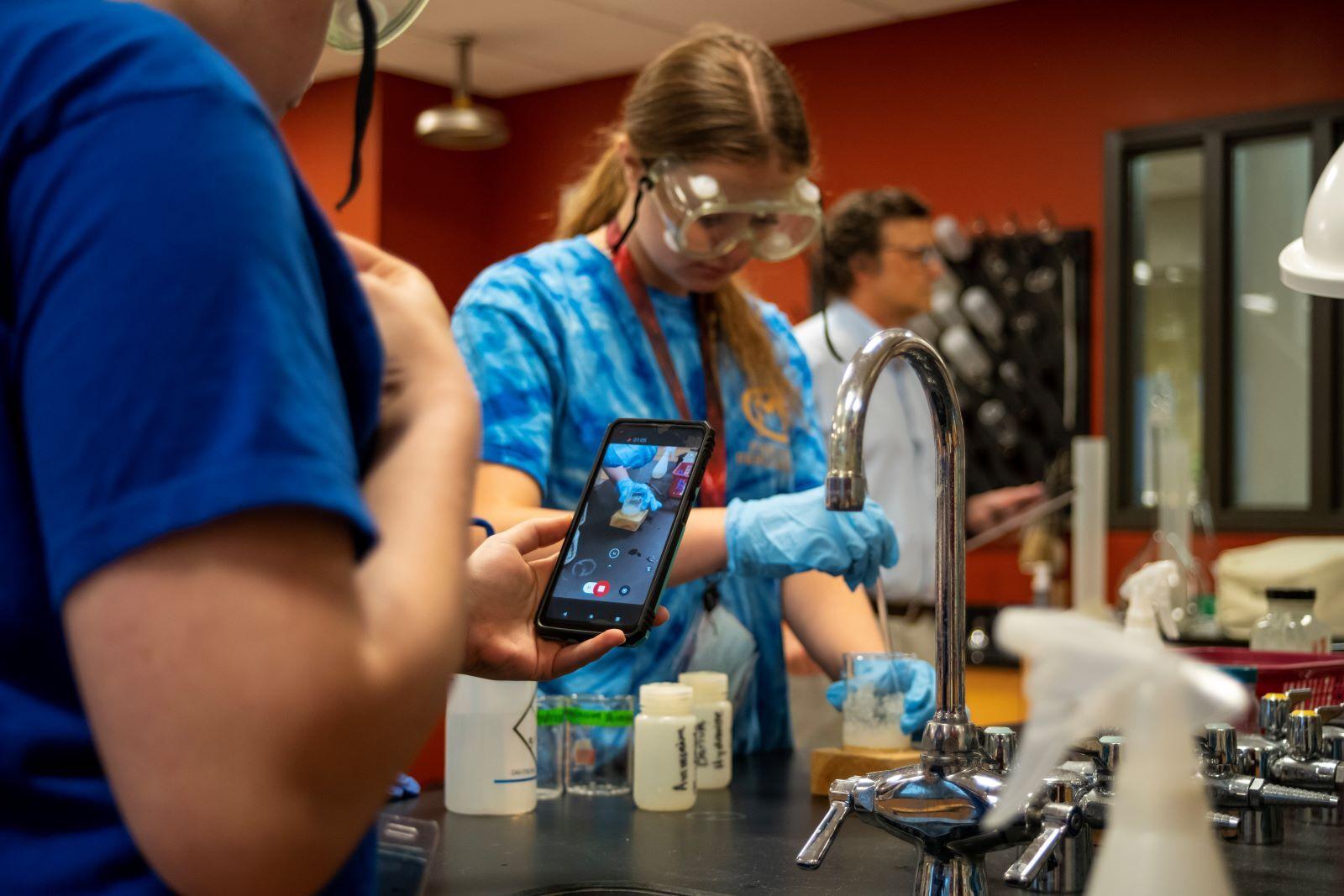 A student wearing blue gloves and goggles takes a video as another student mixes a brown liquid in a glass beaker. The student mixing also wears goggles and a pair of blue gloves. Both students wear blue t-shirts and are standing at a sink. 