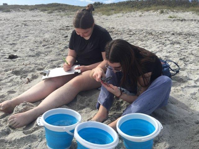 Two students sit in the sand with three blue buckets at their feet. One student is writing on a clip board while the other is looking at something with a special  tool.