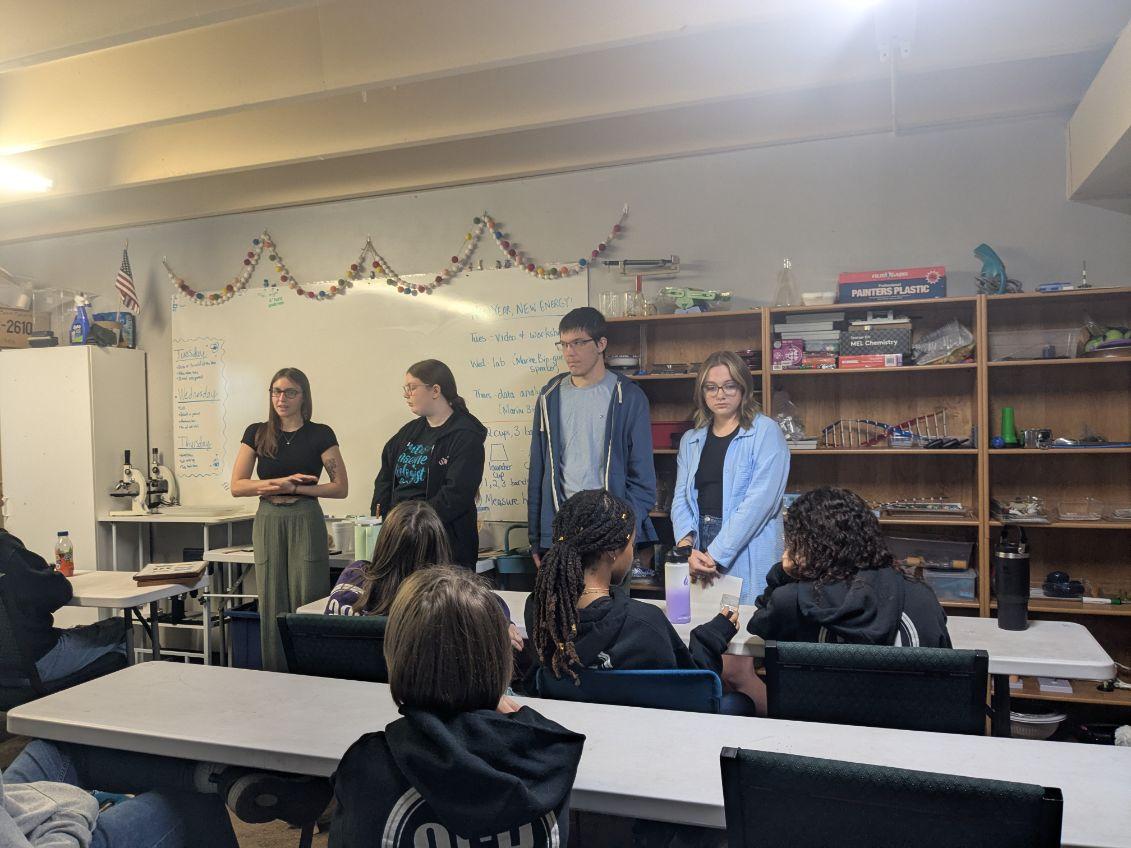 Four students stand in front of a class of children. 