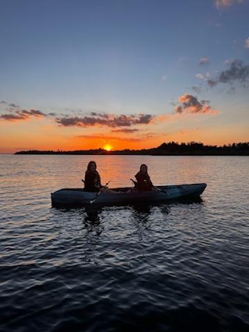 The sky is a deep orange as it sets behind two students sitting in a kayak. 