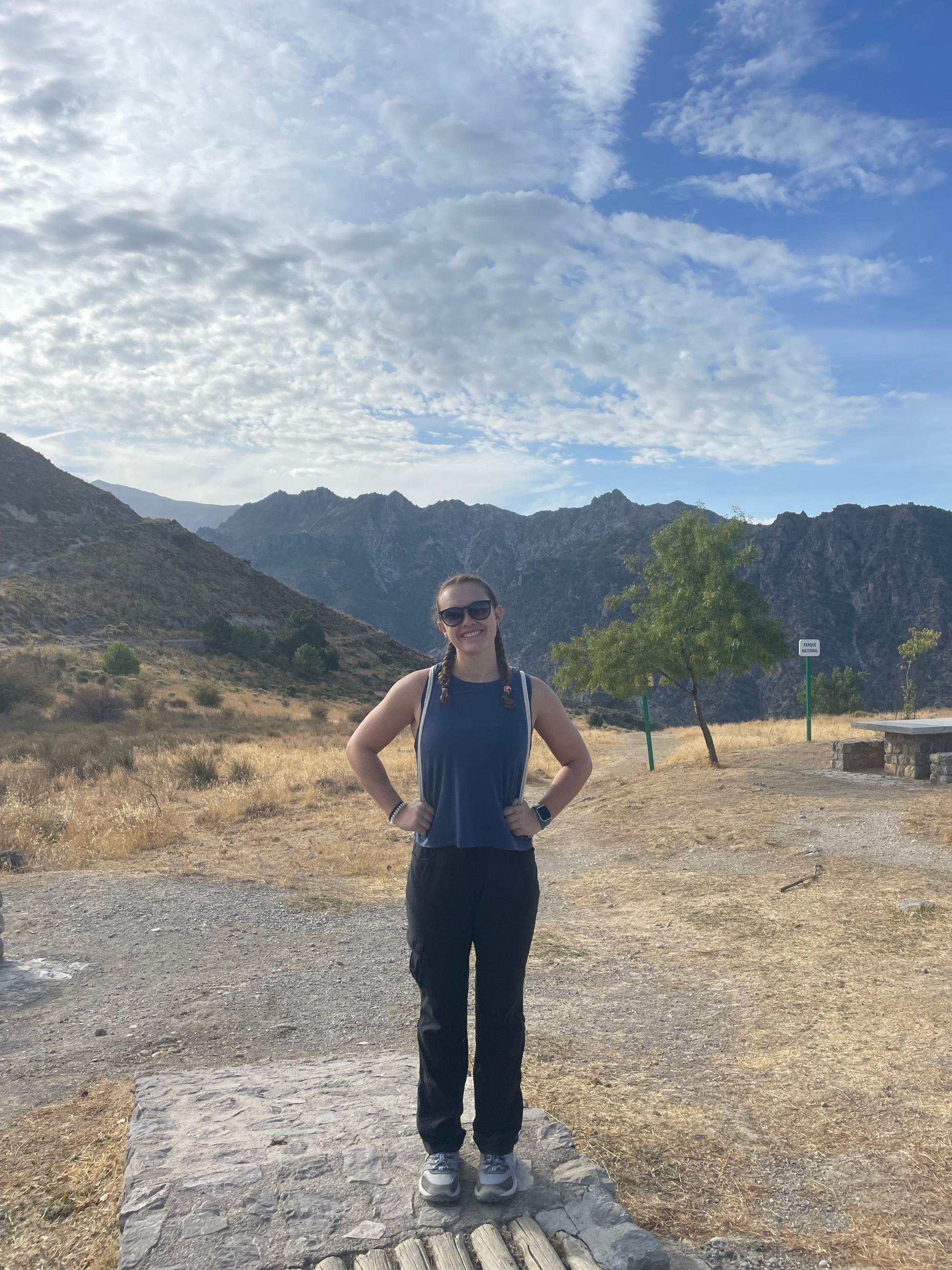 Natalie Double stands in front of hills. She wears a blue tank top and black pants. 