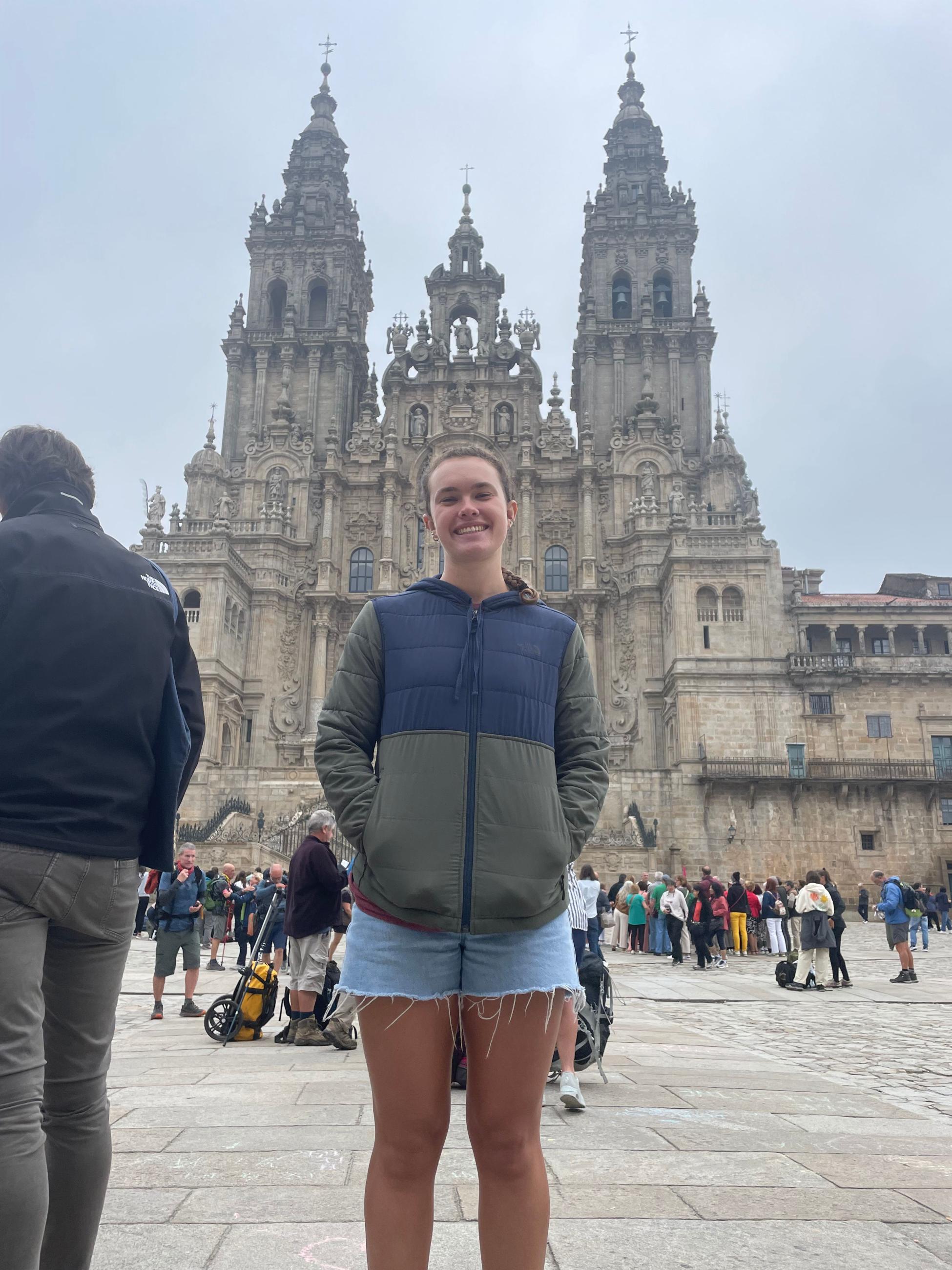 Natalie Double stands in front of the Santiago Cathedral. She wears a blue and green jacket and jean shorts. 