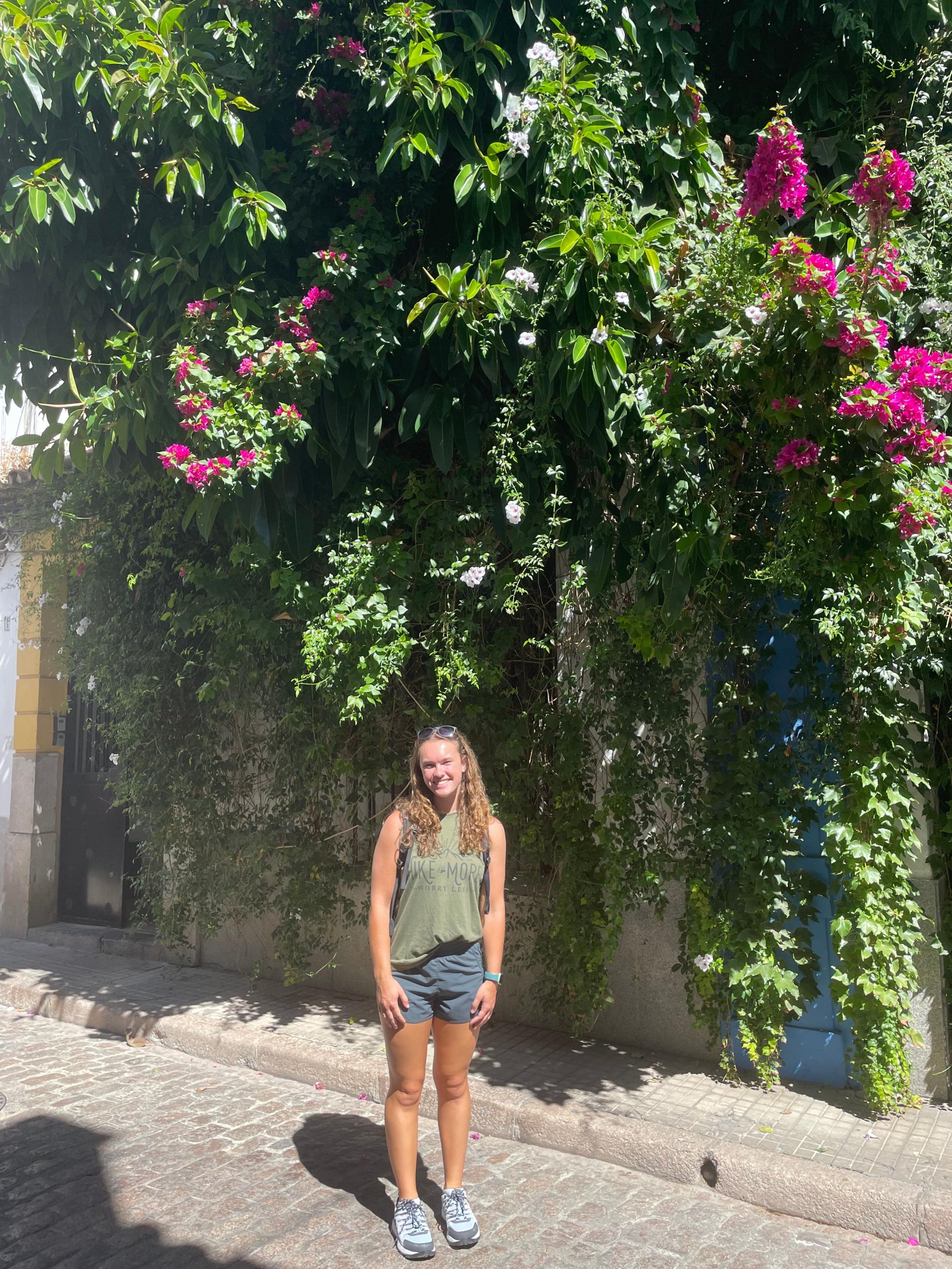 Natalie Double stands in front of a wall of green plants with pink flowers. She wears a green tank top and athletic shorts. 