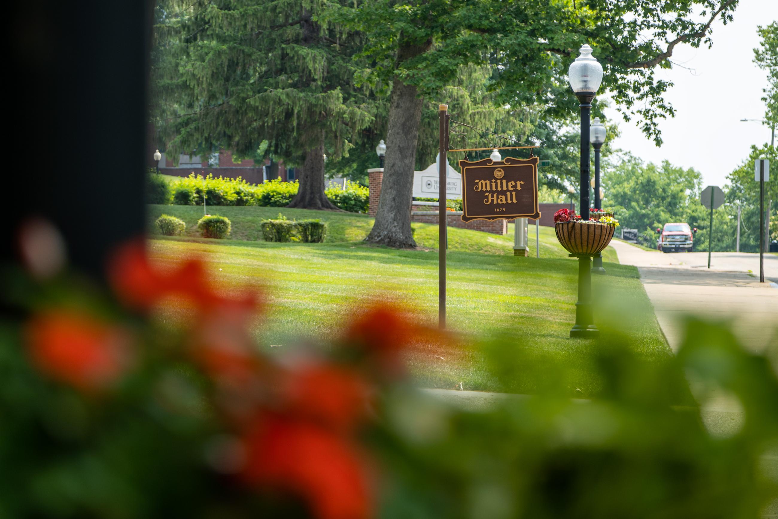 Flowers in the foreground with the Miller Hall and Waynesburg University signs in the background on a sunny day