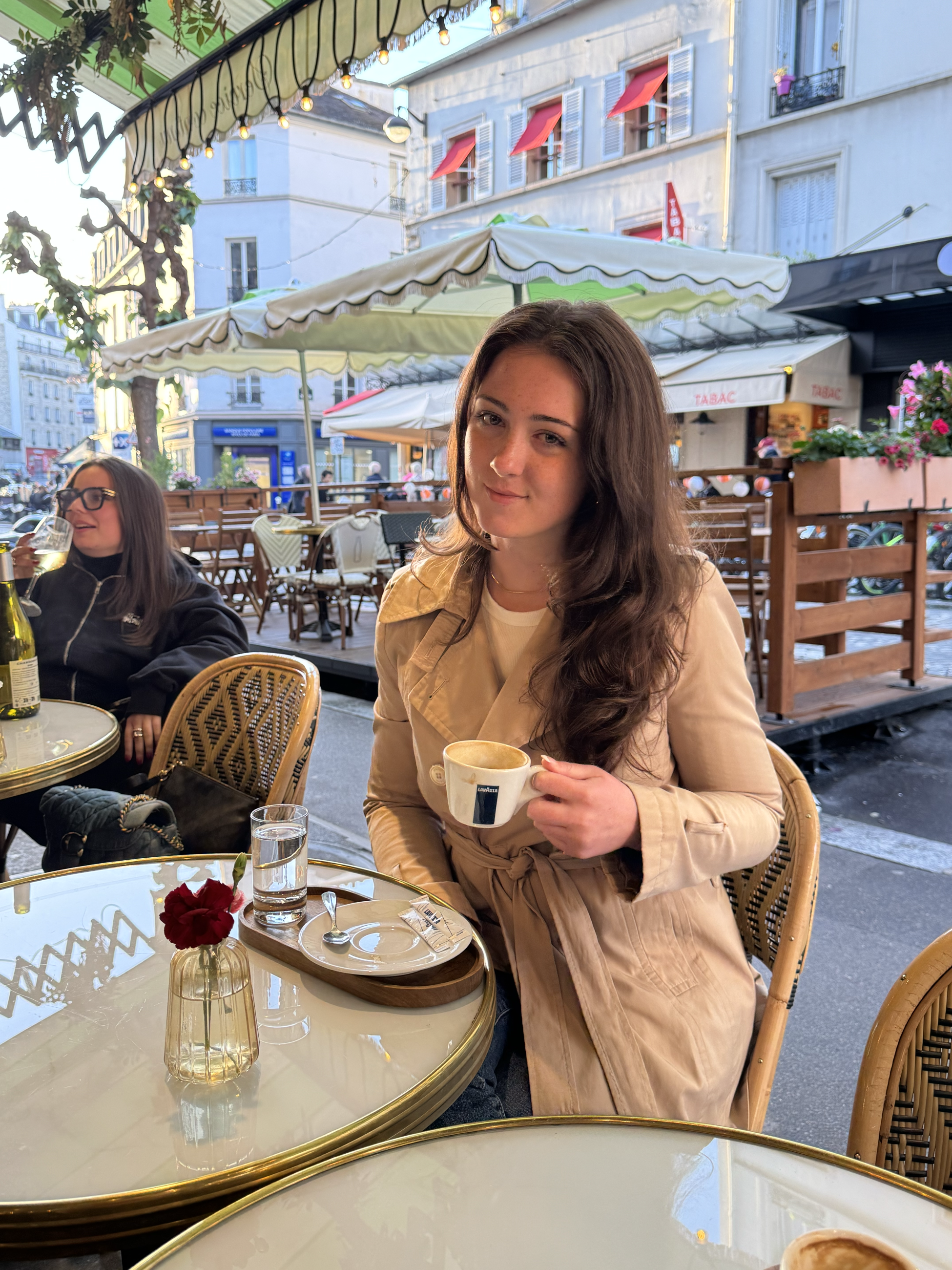 Meredith in a trenchcoat holding a coffee at an outdoor cafe table