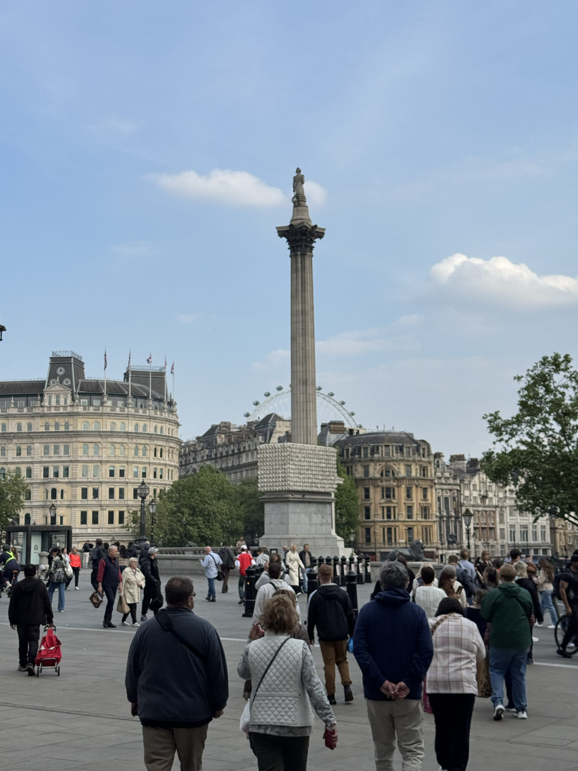 A tall stone column called nelson's Column sits in Trafalgar square, surrounded by decorative pools and buildings.