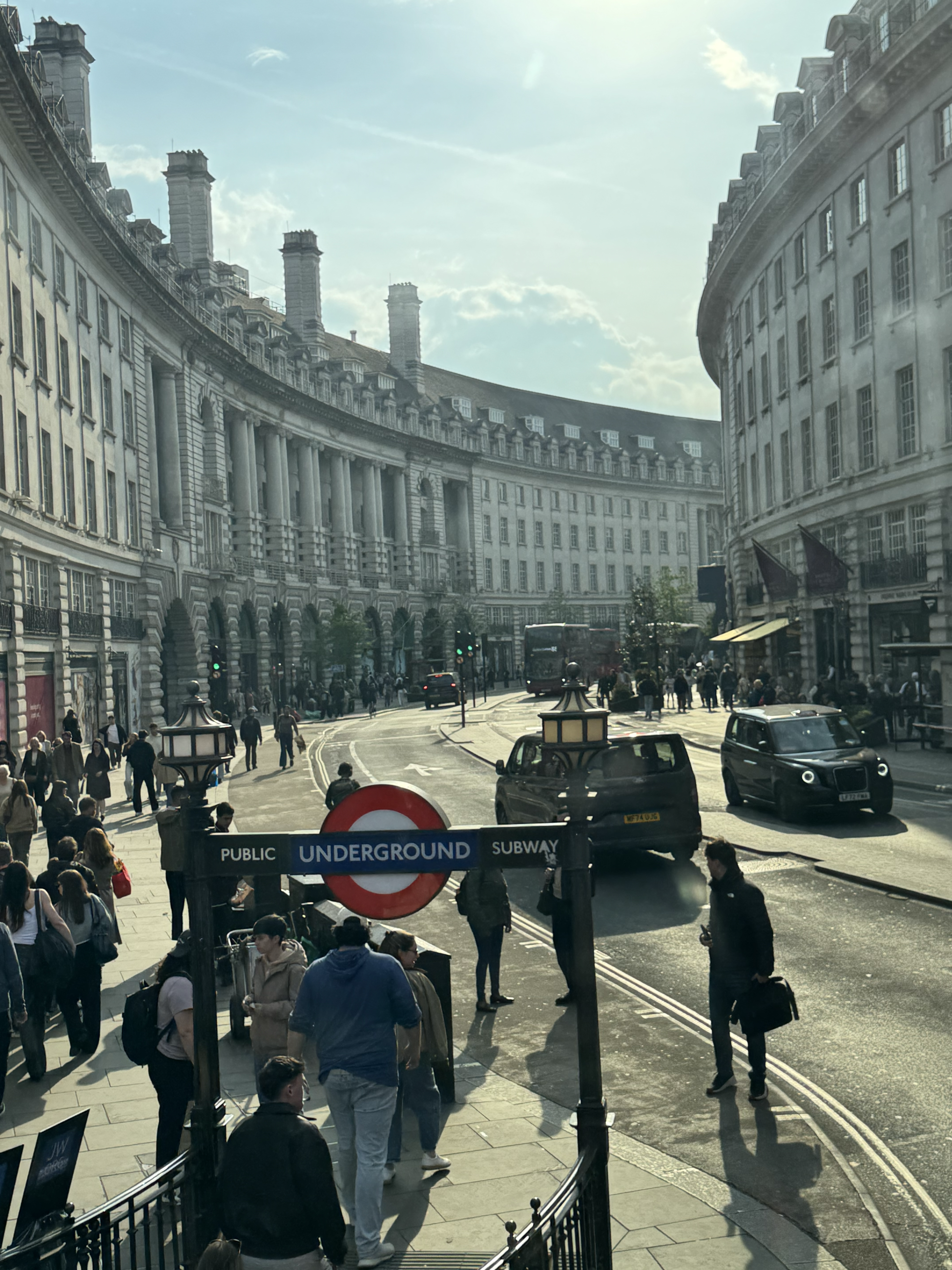 Tall stone buildings lining each side of a curved street, with people entering the underground subway entrance in the foreground. 