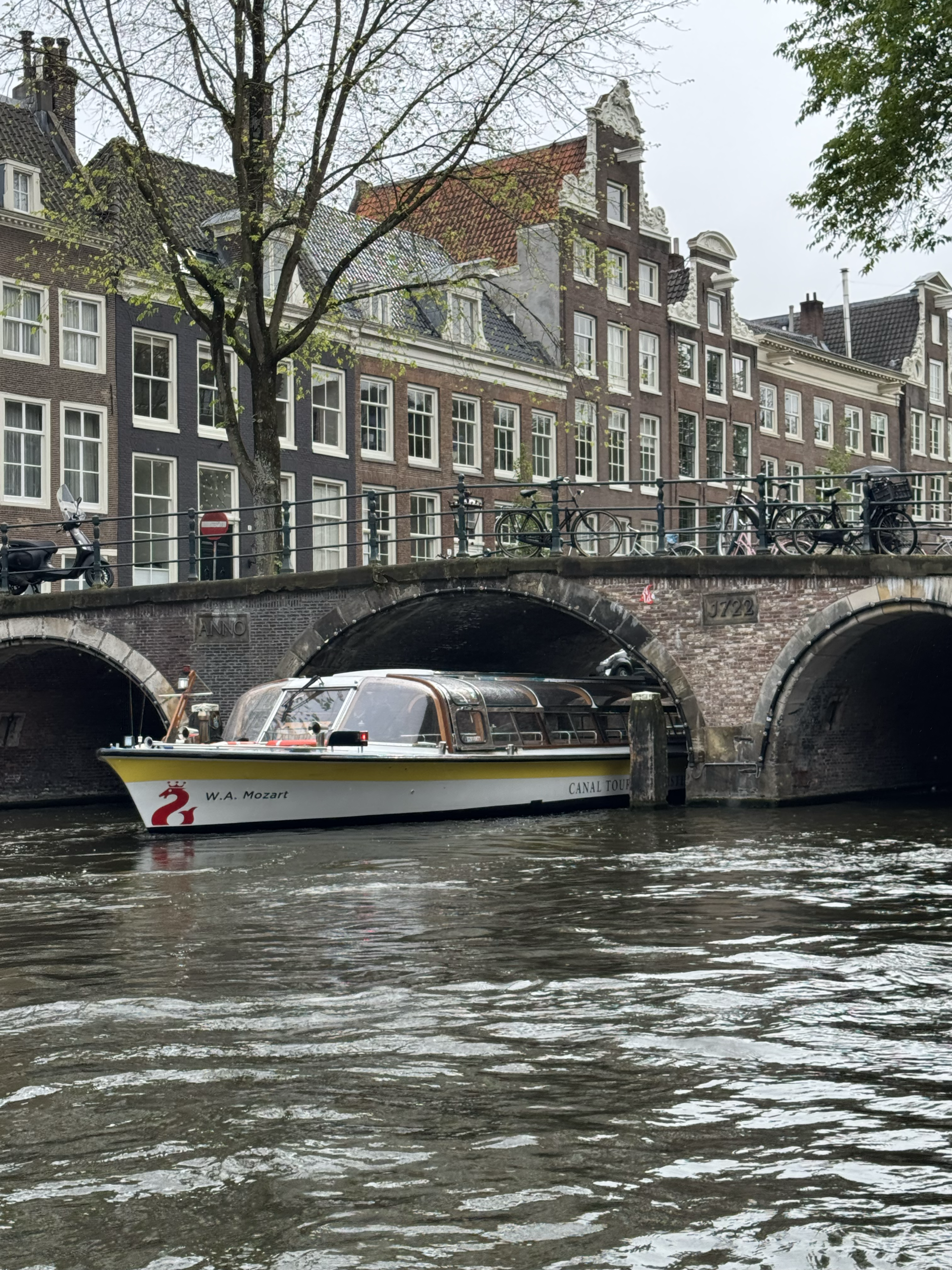 A boat floats under a bridge on a canal, with tall buildings and trees in the background.