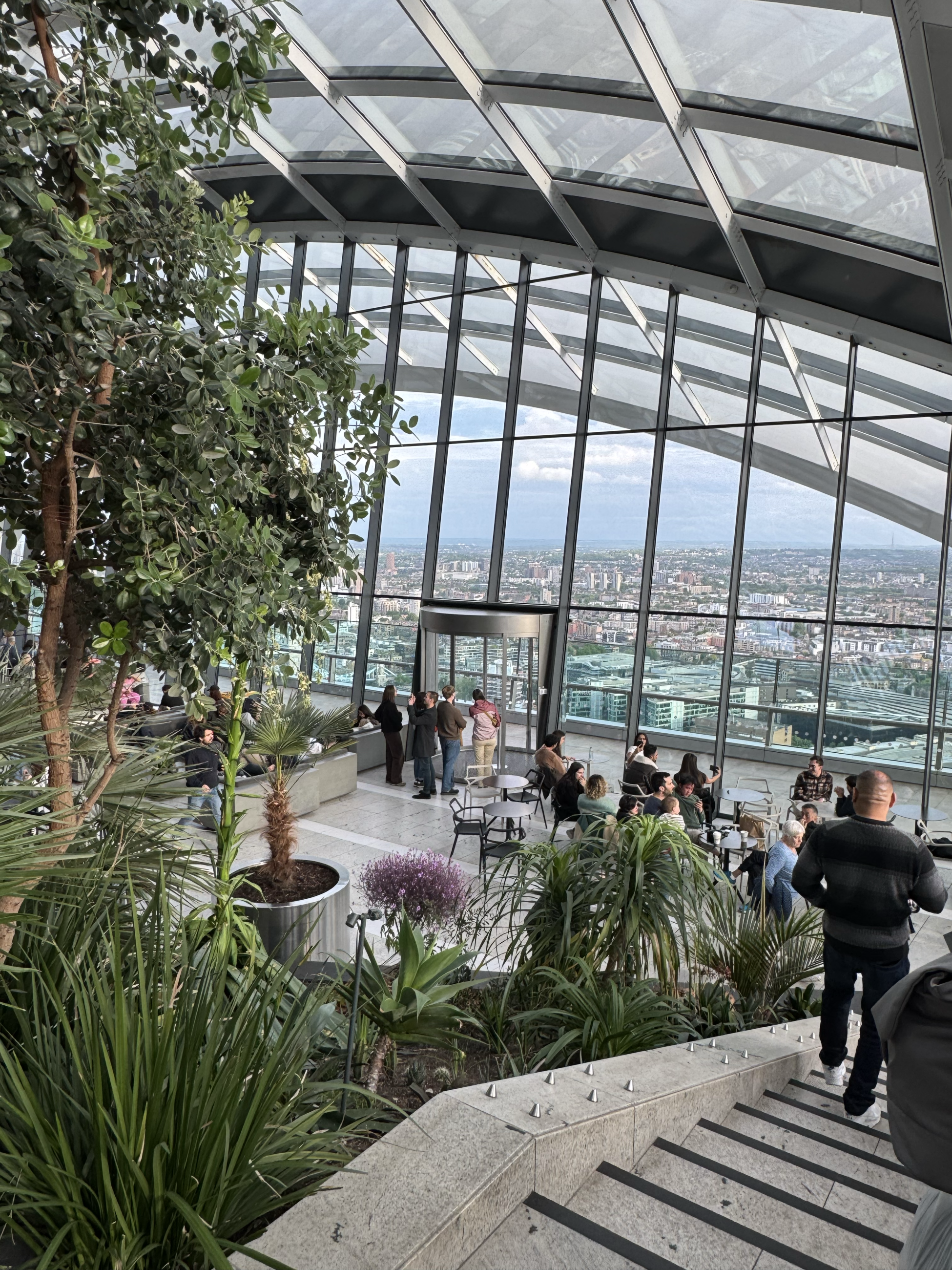 Large glass windows enclose an indoor garden with a variety of trees and plants, and city buildings can be seen through the windows. 
