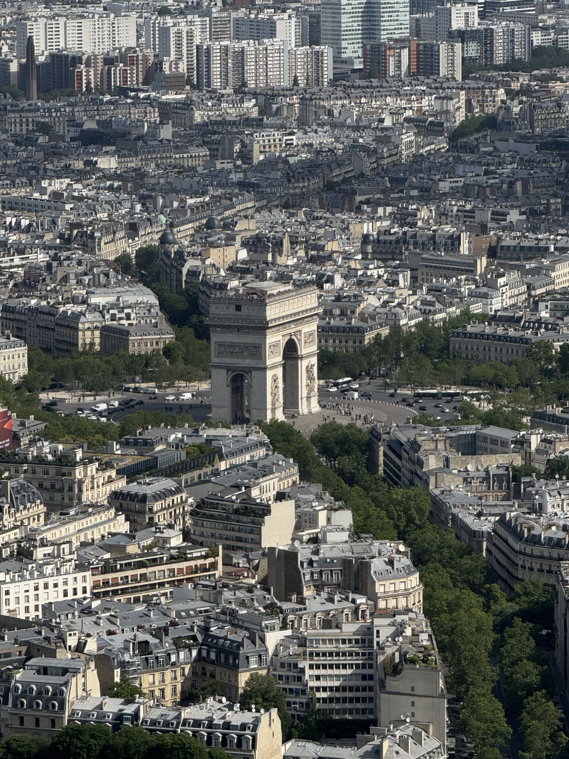 The Arc de Triomphe, a stone arch, sits in the middle of a major intersection, surrounded by trees and buildings. 
