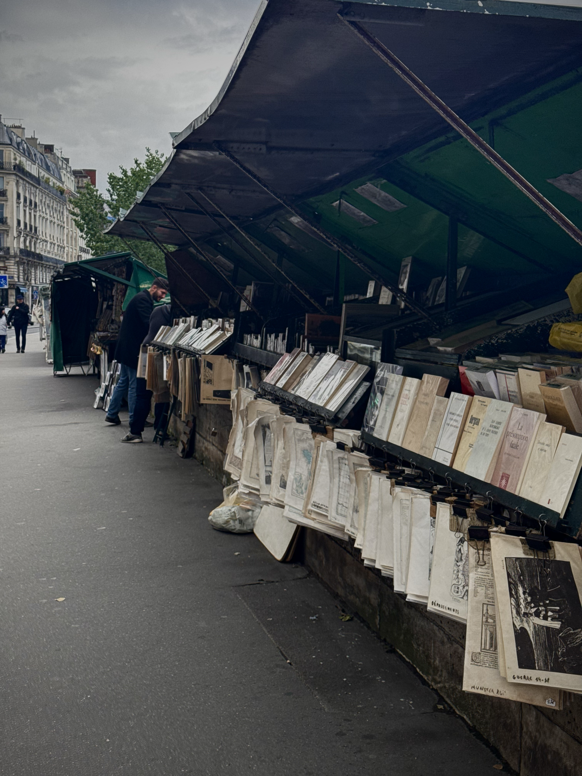 Photography prints and other artwork hangs from outdoor market booths on a Parisian street. Some shoppers can be seen in the distance.