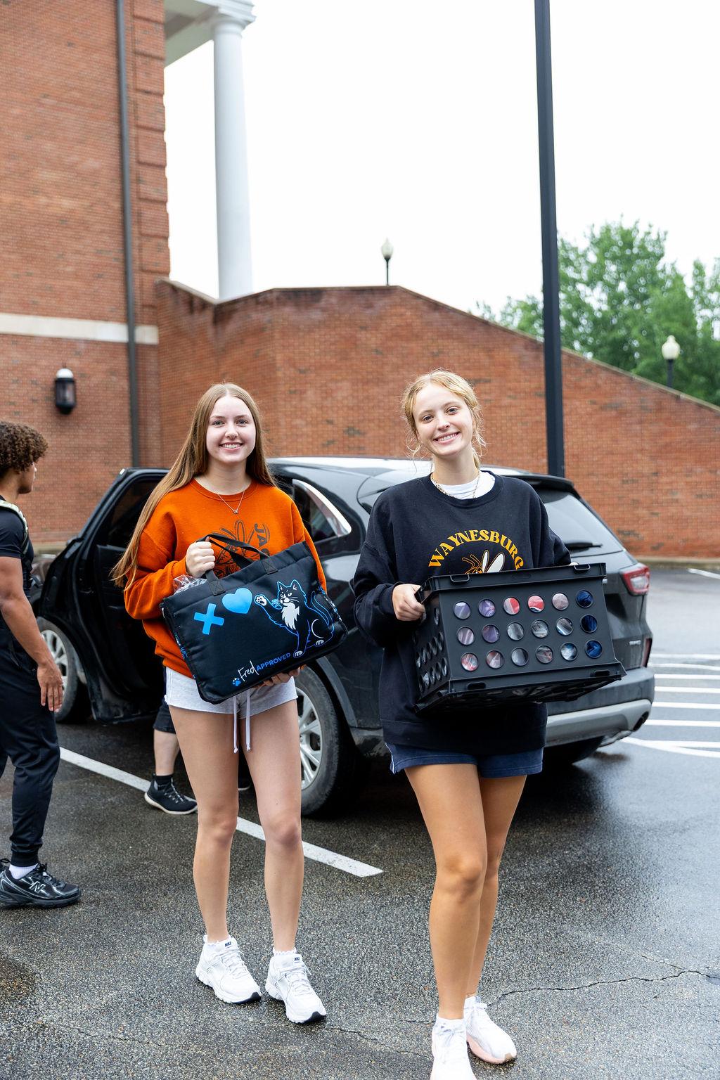 Two students pose for the camera with arms full of dorm items on move in day.