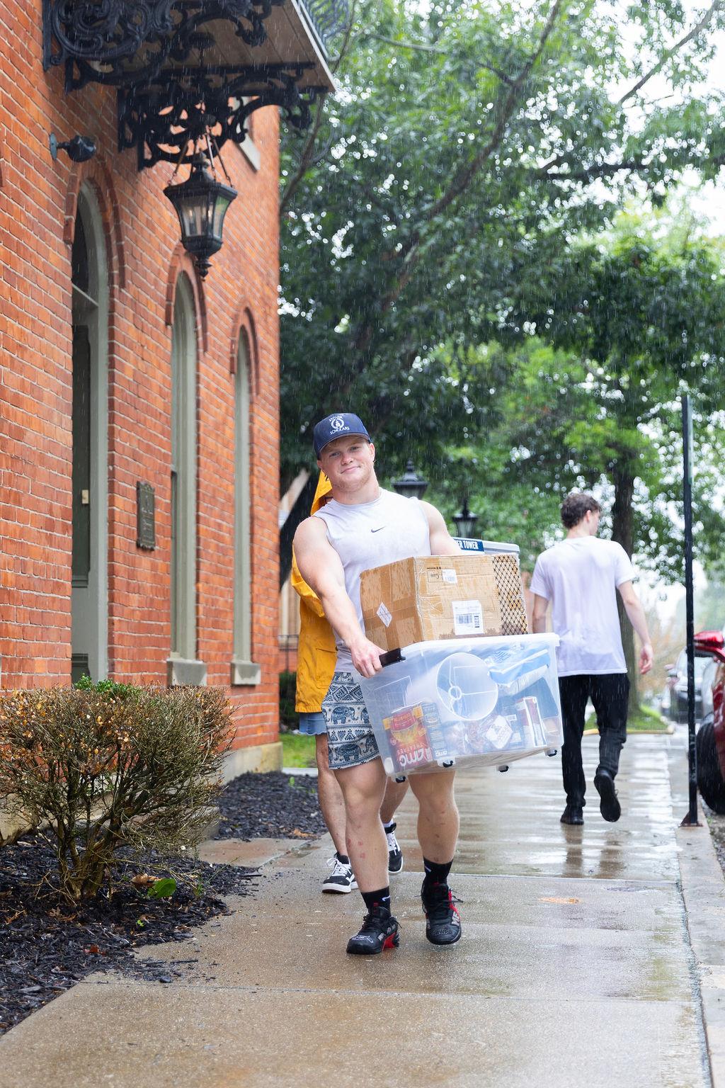 A student caries a tote and box, while walking on a sidewalk, assisting with new student move in.