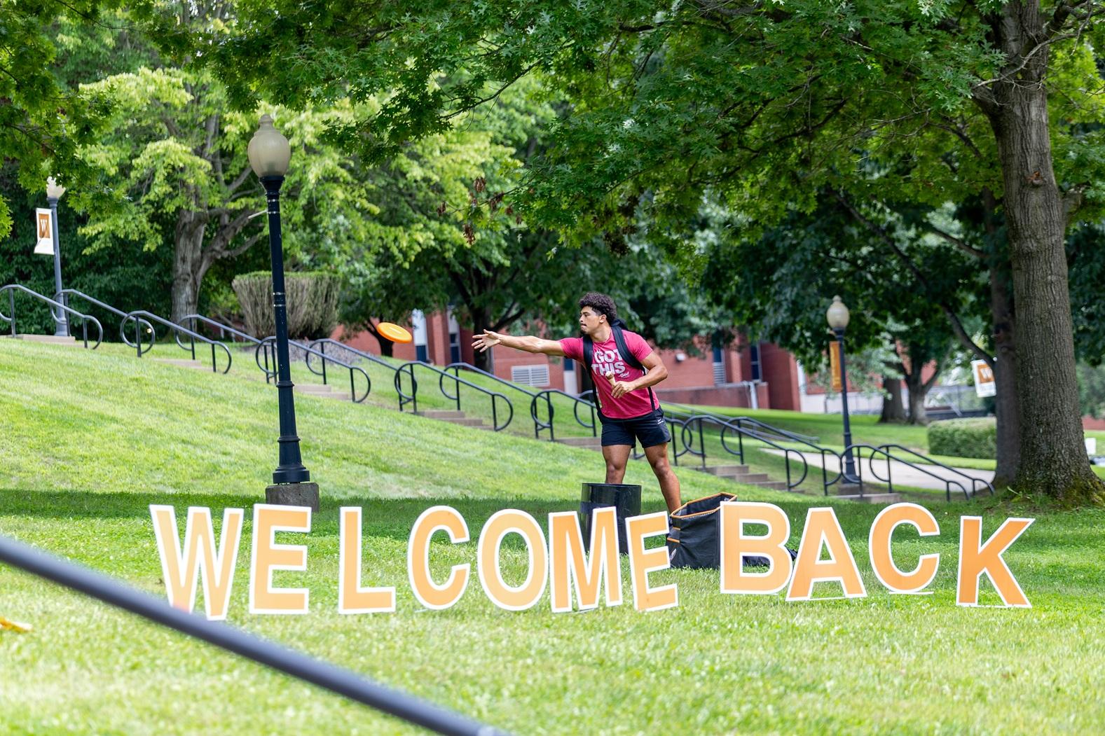 Welcome back sign in the park with a student throwing a frisbee in the background.
