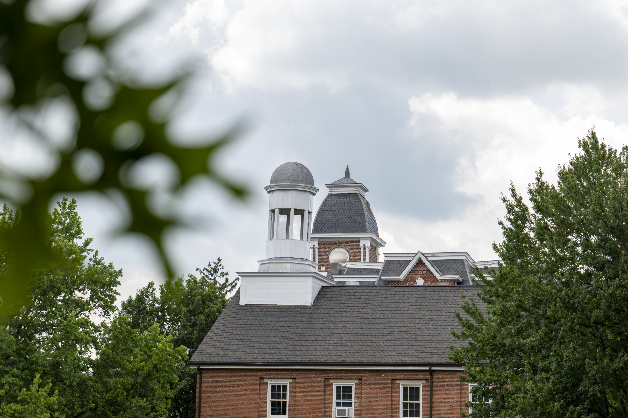 The bell towers of Miller and Hanna hall are in focus against a cloudy sky, with green oak leafs out of focus in the foreground