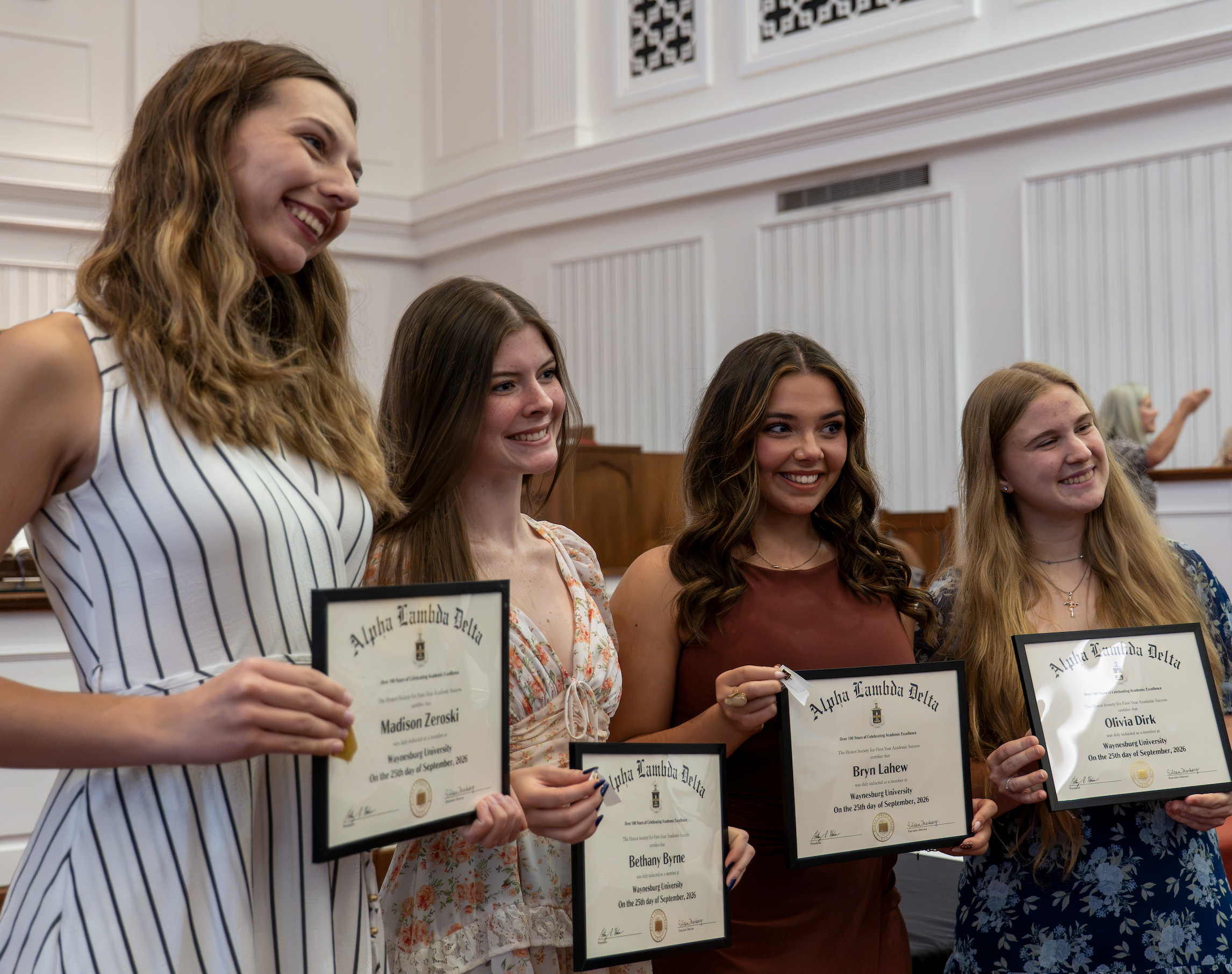 Four students standing together at the front of Roberts Chapel, holding their ALD membership certificates and smiling 