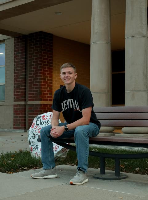 Levi Tracy sits on a brown bench next to a white rock painted with handprints. He wears a black t-shirt and jeans.