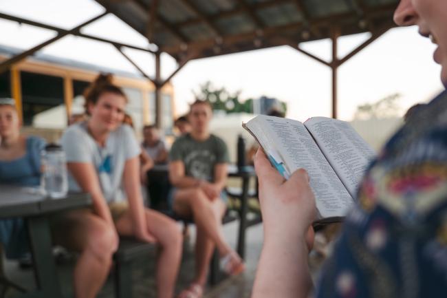 A woman holds a bible open in front of her as she reads. Other group members are seen listening to her.
