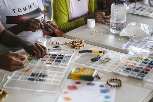 Materials to make bracelets are spread out on a white folding table. Women use beads and string to make colorful bracelets.