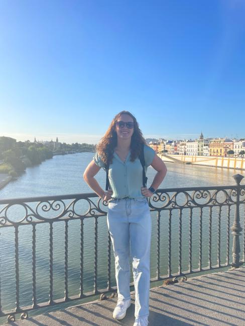 Natalie Double stands on a bridge in front of a river in Spain. She is wearing a green shirt and jeans. 