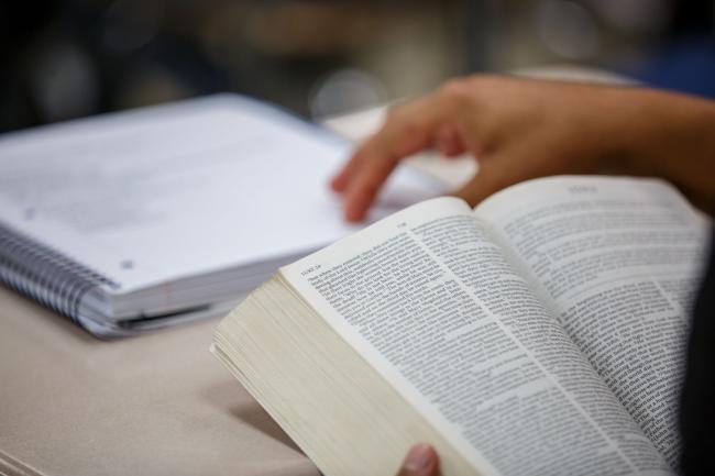 A person holds a bible in front of them with it resting on a table. Their hand is touching a notebook in front of them. 