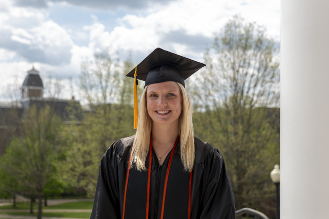 Clara Paige Miller posing in her cap and gown with Miller Hall in the background