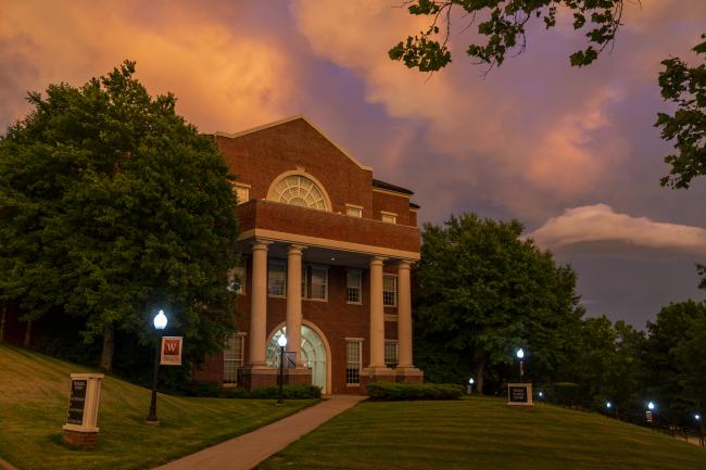 Stover Campus Center at sunset, with an orange and purple sky behind it and the walkways lit by lampposts 
