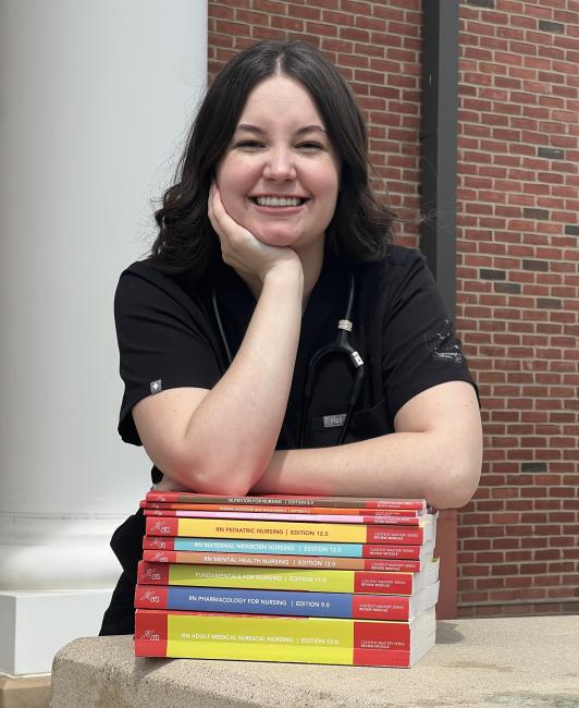A female nursing student in scrubs poses with a stack of nursing textbooks