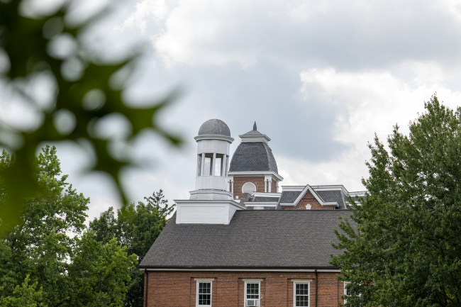The bell towers of Miller and Hanna hall are in focus against a cloudy sky, with green oak leafs out of focus in the foreground