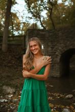 Gracie is pictured in front of a stone bridge with leaves spread around her. She is wearing an emerald green dress and has her hair half up with a white bow. She holds one arm touching her should and the other in front of her.